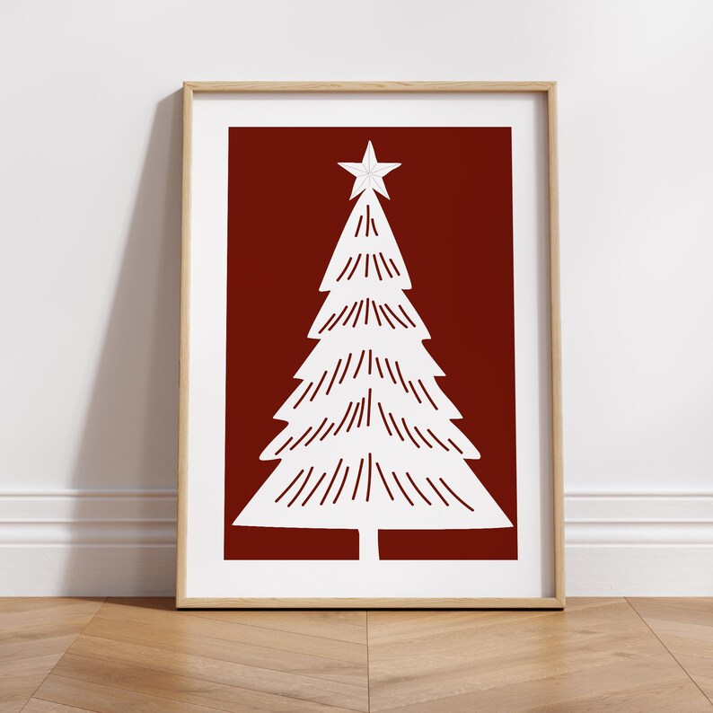 May include: A framed print featuring a white Christmas tree design on a deep red background. The tree is topped with a star and has a minimalist style. The frame is a light wood color, and the print is displayed on a wooden floor.