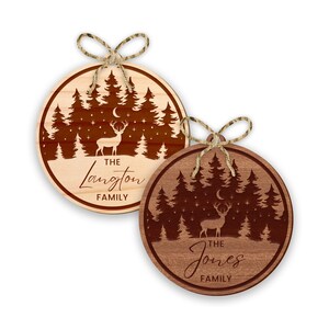 Op de afbeelding: Twee ronde houten ornamenten met een herten silhouet, een halve maan en dennenbomen. De bovenste ornament is van licht hout met een bruin hert en de tekst "THE Langton FAMILY". De onderste ornament is van donker hout met een bruin hert en de tekst "THE Jones FAMILY".