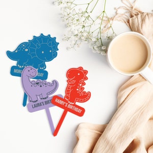May include: Three colorful dinosaur-shaped cake toppers with personalized messages. The blue dinosaur says "Morgan's Day", the purple dinosaur says "Laura's Birthday", and the red dinosaur says "Harry's Birthday".