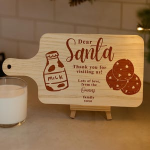 May include: A wooden cutting board with a handle, featuring a milk bottle and cookies illustration. The board is engraved with the words "Dear Santa" and a message from the Owens family. A glass of milk sits to the left.