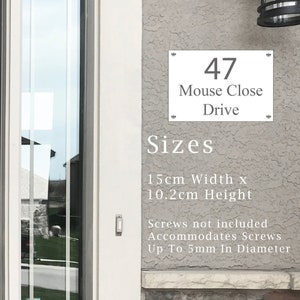 May include: White house number sign with black text, reading "47 Mouse Close Drive". The sign is 15 cm wide and 10.2 cm high.  The sign is mounted on a gray stucco wall.