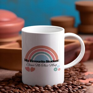 May include: White ceramic mug with a rainbow design and the text "My Favourite Student Gave Me This Mug". The mug is surrounded by coffee beans.