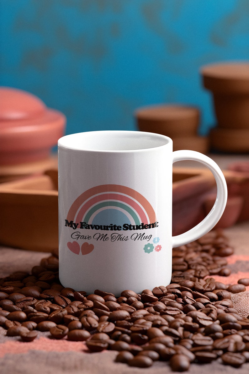 May include: White ceramic mug with a rainbow design and the text "My Favourite Student Gave Me This Mug". The mug is surrounded by coffee beans.
