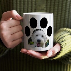 May include: White ceramic mug featuring a black paw print design with a photo of a smiling white dog. The mug is held by a person wearing a green sweater. A fun and unique gift for dog lovers.