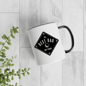 May include: White ceramic mug with a black rim and handle. The mug features a black and white graphic of a diamond with the text "The Best Dad By Par" and a golf ball inside the diamond.