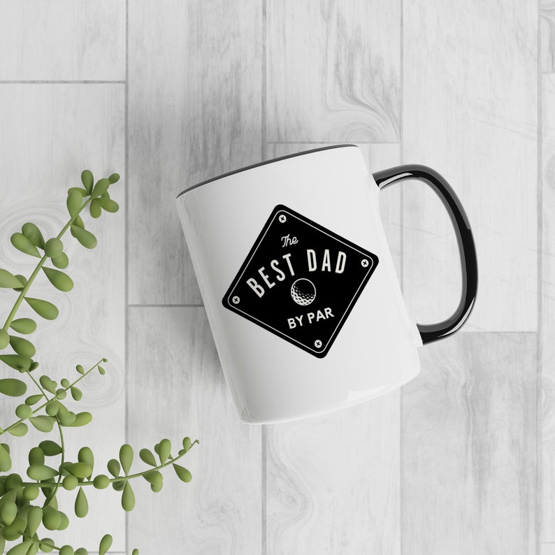 May include: White ceramic mug with a black rim and handle. The mug features a black and white graphic of a diamond with the text "The Best Dad By Par" and a golf ball inside the diamond.