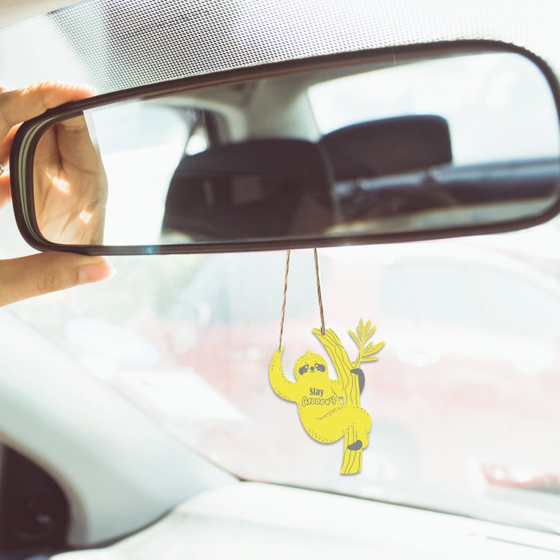 May include: Yellow sloth car air freshener hanging from the rearview mirror of a car. The sloth is holding onto a branch and has the text "Stay groovy" on its belly.