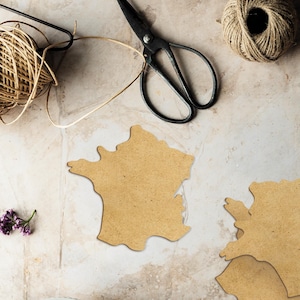 Peut inclure: Vue a&eacute;rienne d'articles de bricolage : une pelote de ficelle, des ciseaux anciens et des d&eacute;coupes en bois en forme de France. Une petite branche de fleurs violettes est &eacute;galement pr&eacute;sente, sugg&eacute;rant un th&egrave;me artisanal ou de bricolage.