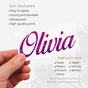 May include: A white sticker with the name "Olivia" in a vibrant purple script font. The sticker is held by a hand and is designed for application on various surfaces, including glass, metal, and plastic. The key features include being easy to apply, strong, durable, waterproof, and having a high-quality print.