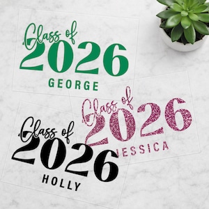 May include: Three clear acrylic graduation announcements with the text "Class of 2026" and a name below. One is green, one is black, and one is glittery pink. A small succulent plant in a white pot is in the background.
