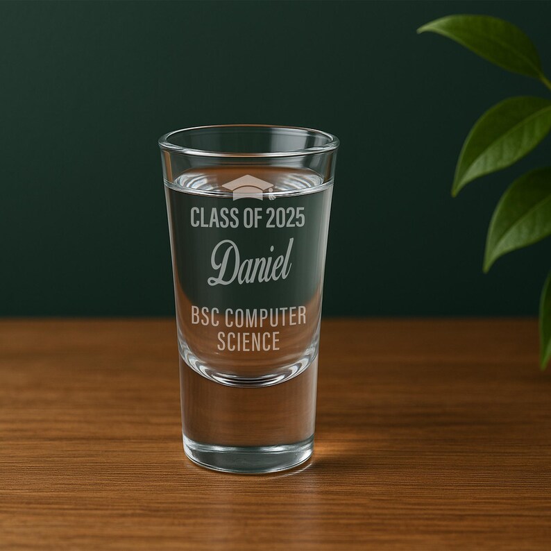 May include: A clear shot glass filled with liquid, featuring etched text: "CLASS OF 2025", "Daniel", and "BSC COMPUTER SCIENCE". The glass is on a wooden surface with a dark green background and a plant in the corner.