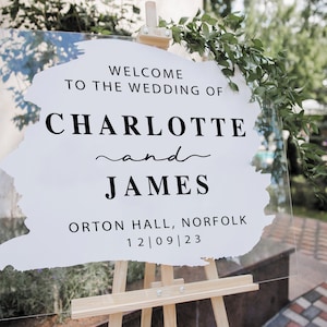 May include: A clear acrylic wedding welcome sign on a wooden easel. The sign features the text "WELCOME TO THE WEDDING OF CHARLOTTE and JAMES" in black, along with the location and date. Greenery decorates the top.