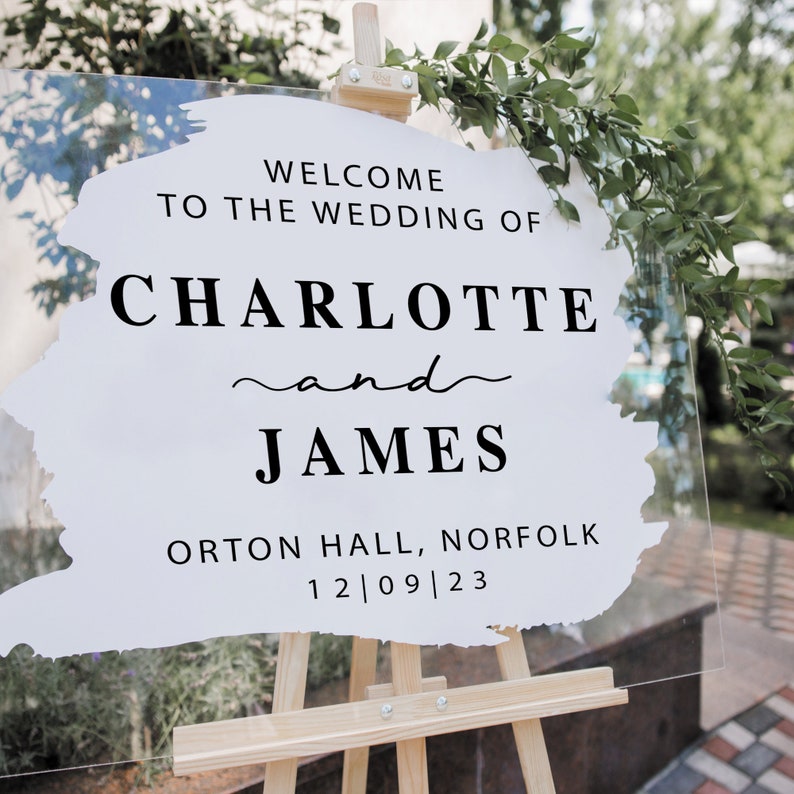 May include: A clear acrylic wedding welcome sign on a wooden easel. The sign features the text "WELCOME TO THE WEDDING OF CHARLOTTE and JAMES" in black, along with the location and date. Greenery decorates the top.
