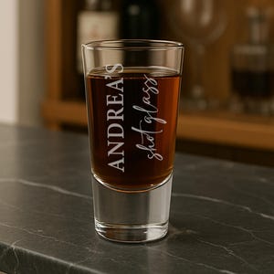 May include: A clear shot glass filled with a dark amber liquid. The glass is engraved with the name "Andrea's" and the words "shot glass" in a cursive font. The glass sits on a dark gray surface.