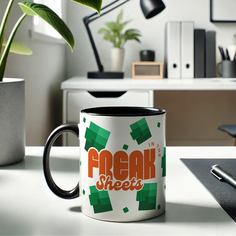 May include: A white ceramic mug with a black rim and handle. The mug has a green and white pixelated design and the text "Freak Sheets" in orange. The text "In the" is in white and is smaller than the other text.