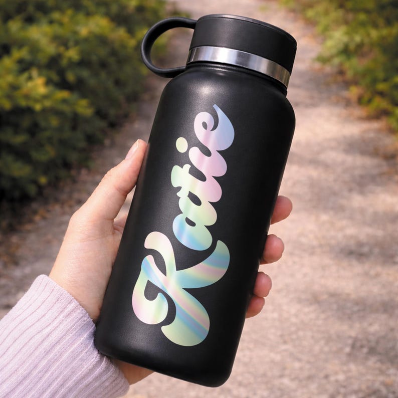 Puede incluir: Una botella de agua aislada negra con una banda plateada y un asa negra. La palabra "Katie" est&aacute; escrita en una escritura hologr&aacute;fica de color pastel en la parte delantera de la botella. La botella se sostiene al aire libre.