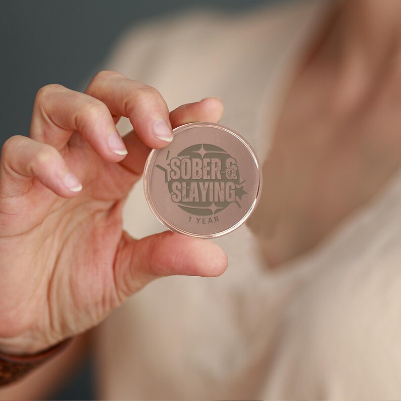 Puede incluir: Una moneda de sobriedad redonda, transparente y de pl&aacute;stico con el texto "SOBER & SLAYING" y "1 YEAR" impreso en ella.
