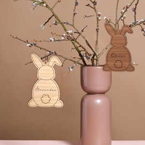 May include: Two wooden bunny ornaments with names "Alexandre" and "Oliver" engraved on them. The ornaments are hanging from a branch of a pussy willow tree in a brown vase.