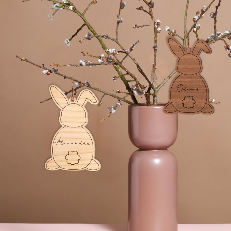 May include: Two wooden bunny ornaments with names "Alexandre" and "Oliver" engraved on them. The ornaments are hanging from a branch of a pussy willow tree in a brown vase.