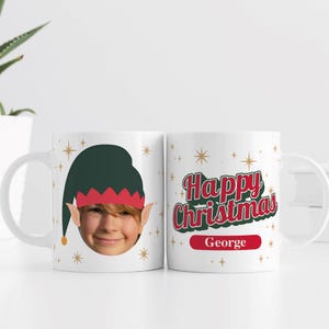 May include: White ceramic mug with a child's face in an elf hat. The other side of the mug says "Happy Christmas" in red and green with the name "George" below. Gold star accents are scattered across the mug.