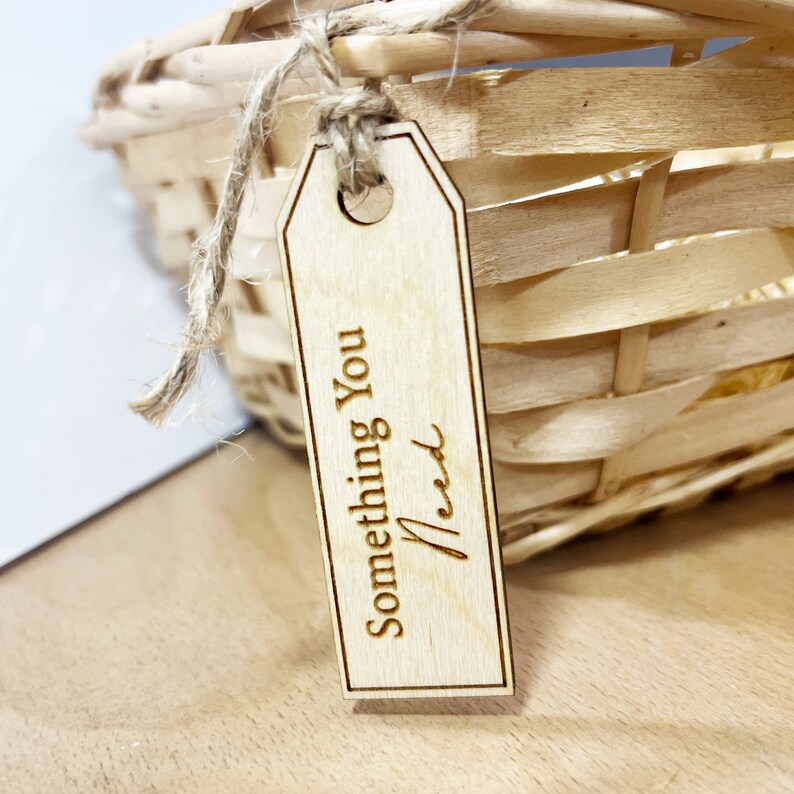 Pu&ograve; includere: Un'etichetta regalo in legno con il testo "Something You Need" inciso su di essa. L'etichetta &egrave; legata con uno spago e pende da un cesto di vimini.