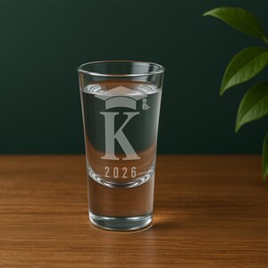 K&ouml;nnte beinhalten: Ein klares Schnapsglas, gef&uuml;llt mit Fl&uuml;ssigkeit, mit einem Abschlusskappen-Design &uuml;ber dem Buchstaben "K" und der Jahreszahl "2026" graviert. Das Glas steht auf einer Holzoberfl&auml;che vor einem dunkelgr&uuml;nen Hintergrund.