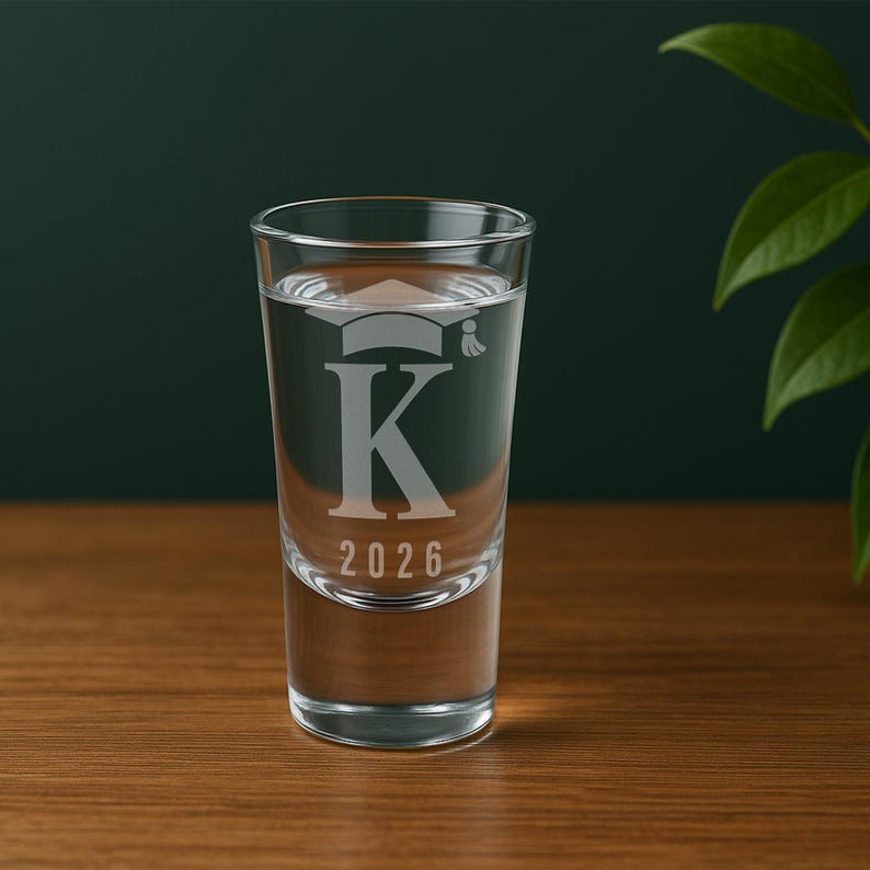 K&ouml;nnte beinhalten: Ein klares Schnapsglas, gef&uuml;llt mit Fl&uuml;ssigkeit, mit einem Abschlusskappen-Design &uuml;ber dem Buchstaben "K" und der Jahreszahl "2026" graviert. Das Glas steht auf einer Holzoberfl&auml;che vor einem dunkelgr&uuml;nen Hintergrund.