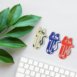 May include: Three cat-shaped bookmark designs. The bookmarks are made of paper and are in the shape of a cat. The cat designs are a black and white cat, a blue and white cat, and a red and white cat. The bookmarks are on a white surface.
