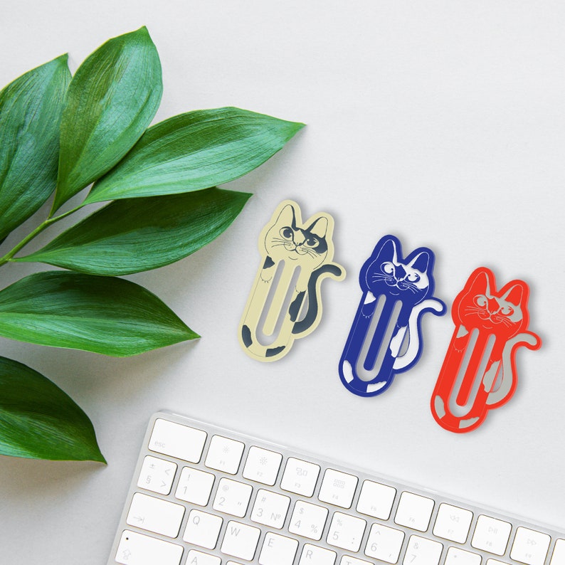 May include: Three cat-shaped bookmark designs. The bookmarks are made of paper and are in the shape of a cat. The cat designs are a black and white cat, a blue and white cat, and a red and white cat. The bookmarks are on a white surface.