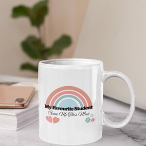 May include: White ceramic mug with a rainbow design and the text "My Favourite Student Gave Me This Mug". The mug has two pink hearts and two blue flowers on the bottom.