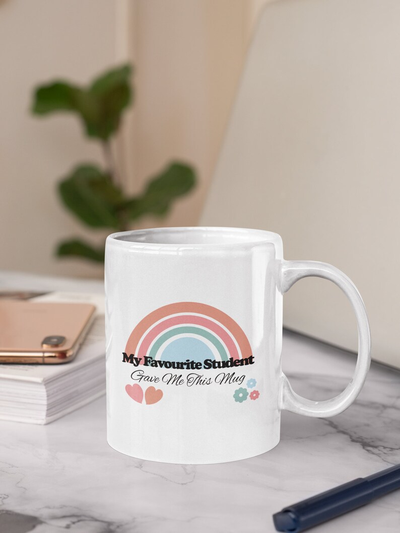 May include: White ceramic mug with a rainbow design and the text "My Favourite Student Gave Me This Mug". The mug has two pink hearts and two blue flowers on the bottom.