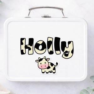 May include: White metal lunchbox with a silver handle. The name "HOLLY" is printed in large, black and white cow-print letters. A cartoon cow sticker with black spots and a pink face is below the name.