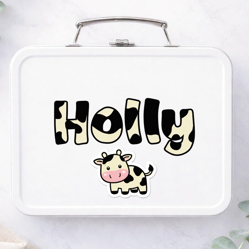 May include: White metal lunchbox with a silver handle. The name "HOLLY" is printed in large, black and white cow-print letters. A cartoon cow sticker with black spots and a pink face is below the name.