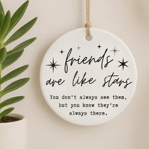 May include: A white ceramic disc with the text "friends are like stars" in black script. The disc has a string for hanging and is decorated with starburst designs. The quote continues: "You don't always see them, but you know they're always there."