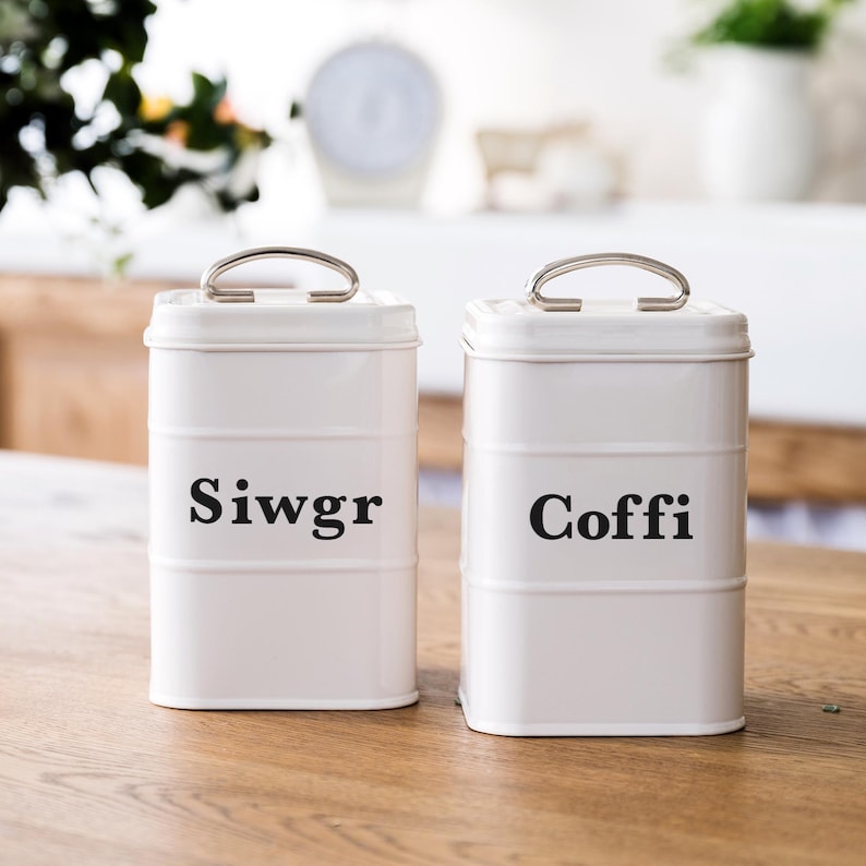 May include: Two white metal canisters with silver handles. One canister reads "Siwgr" in black text, and the other reads "Coffi". The canisters are cylindrical with horizontal bands and sit on a wooden surface.