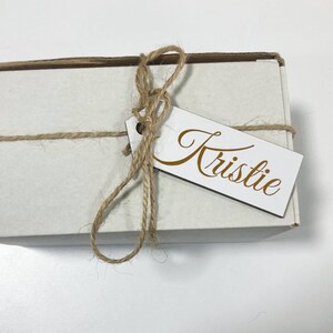 May include: A white gift box tied with twine and a wooden gift tag with the name "Kristie" written in gold cursive.