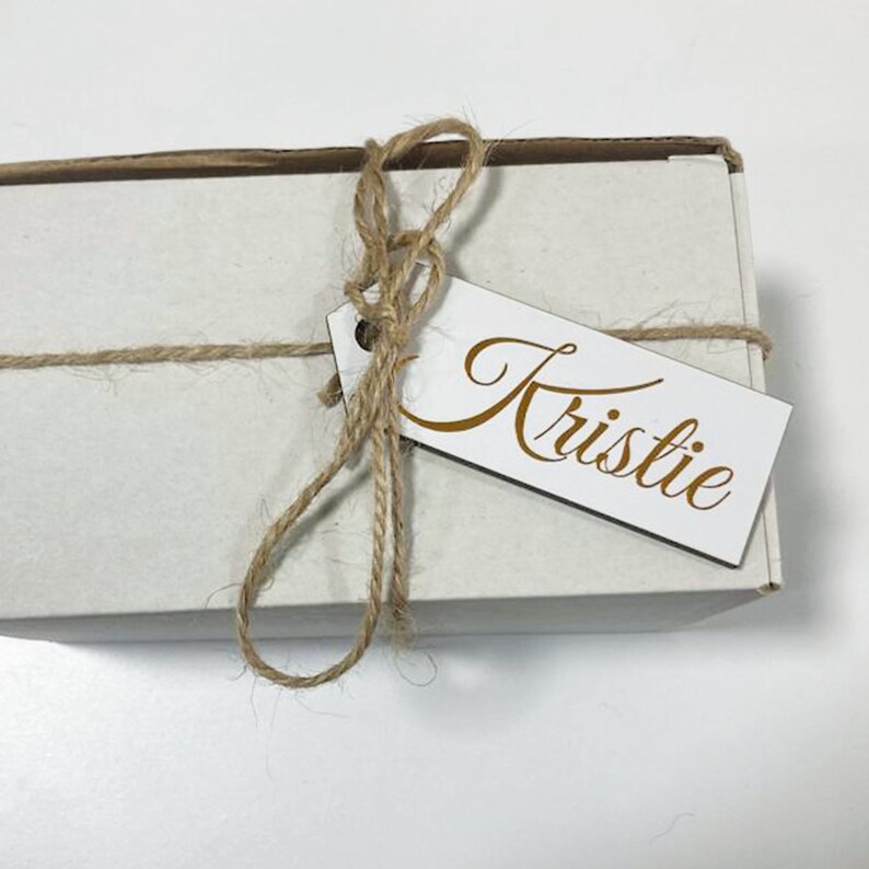 May include: A white gift box tied with twine and a wooden gift tag with the name "Kristie" written in gold cursive.