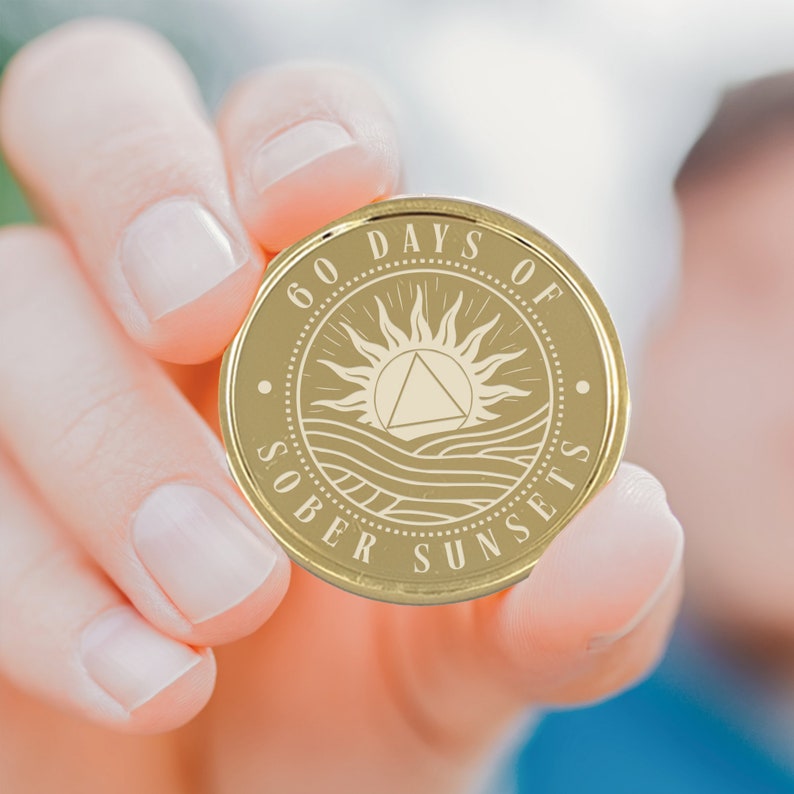 May include: Gold-colored coin with the words "60 Days of Sober Sunsets" and a sun with a triangle inside, surrounded by waves.