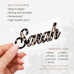 May include: A clear sticker with the name "Sarah" in a leopard print design. The sticker is held by a hand and is suitable for glass, plastic, metal, mirrors, smooth surfaces, and bottles. Key features include easy application, durability, and a waterproof design.