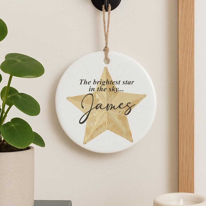 May include: A round, white ceramic ornament with a gold star design and the name "James" in cursive. The text "The brightest star in the sky..." is above the star. A jute string is attached for hanging.
