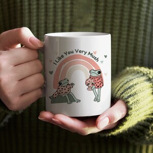 Puede incluir: Taza de cer&aacute;mica blanca con un arco&iacute;ris y dos ranas que llevan jers&eacute;is. El texto en la taza dice "I Like You Very Mush".