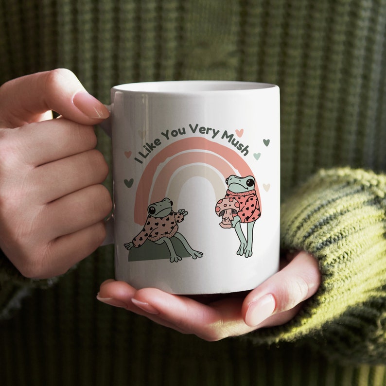 Puede incluir: Taza de cer&aacute;mica blanca con un arco&iacute;ris y dos ranas que llevan jers&eacute;is. El texto en la taza dice "I Like You Very Mush".