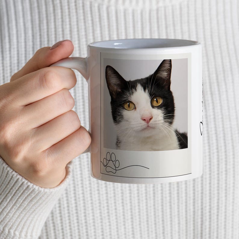 May include: White ceramic mug featuring a black and white cat portrait with yellow eyes. A paw print and curved line are at the bottom of the image. The mug has a white handle.