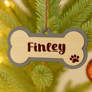 May include: A dog bone-shaped Christmas ornament with the name "Finley" in dark red script. The ornament has a light wood center, a gray border, and a paw print. It hangs from a natural twine string.