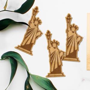 May include: Three wooden cutouts of the Statue of Liberty, each with a brown, textured finish. The cutouts are arranged in a triangular shape on a white surface.