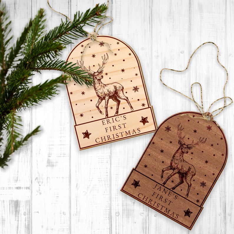 Op de afbeelding: Twee houten kerstversieringen in de vorm van sneeuwbollen met een hert erin. De versieringen zijn gepersonaliseerd met de tekst "Eric's First Christmas" en "Jane's First Christmas".