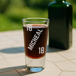 May include: A shot glass filled with a dark liquid, with the name "MICHEAL" and the number "18" etched on it. A dark green bottle is blurred in the background. The image suggests a beverage or bar setting, possibly for a celebration.