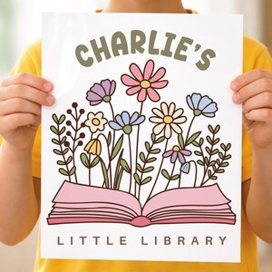 May include: A white print featuring the text "CHARLIE'S" above an open pink book with colorful flowers. The words "LITTLE LIBRARY" are below the book. The flowers include pink, purple, blue, yellow, and white blooms.