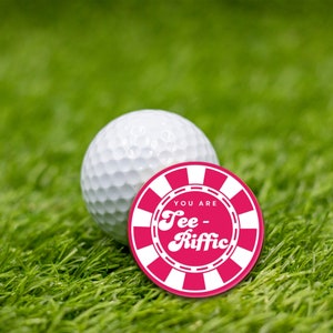 May include: A white golf ball with a pink and white circular sticker that says "You are Tee-Rific" on it.