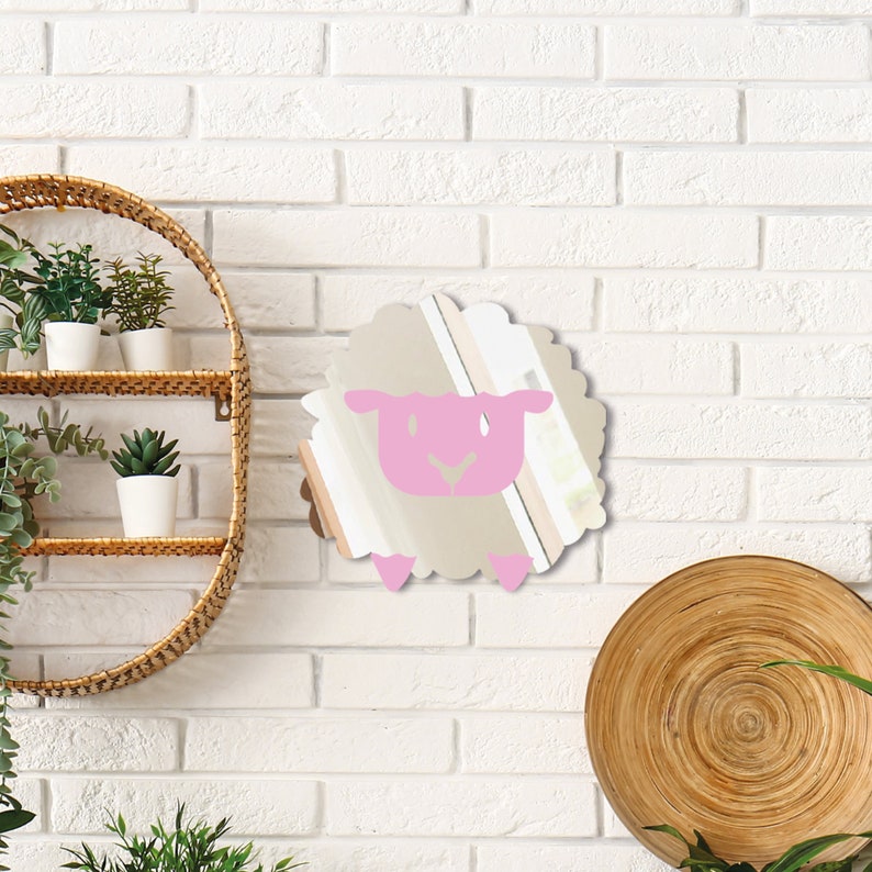 May include: A sheep-shaped mirror with a pink face and feet, mounted on a white brick wall. The mirror has a beige body and a scalloped edge. A woven shelf with plants and a wooden plate are also visible.